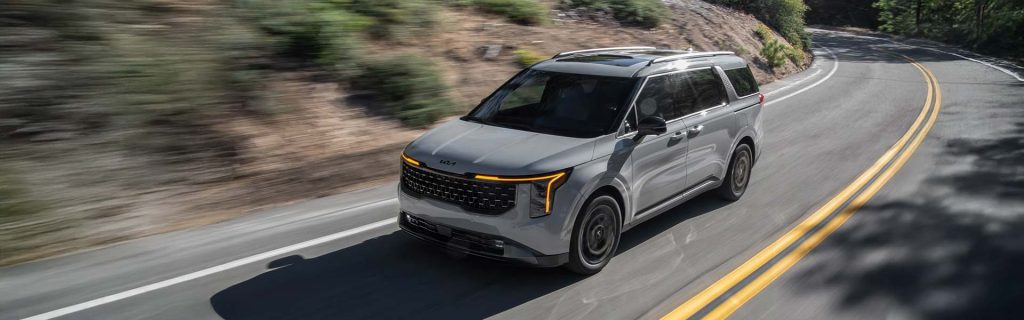 A silver Kia Carnival minivan driving on a curved road.