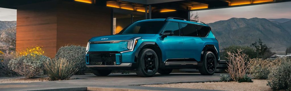 A blue Kia EV9 electric SUV parked outside a modern building with mountains in the background.