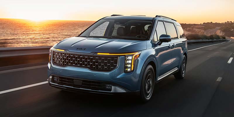 Blue Kia SUV driving along a coastal road at sunset.